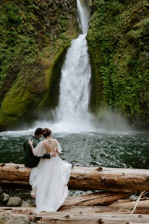 A couple sits on a fallen log admiring a dramatic waterfall in a lush green gorge.