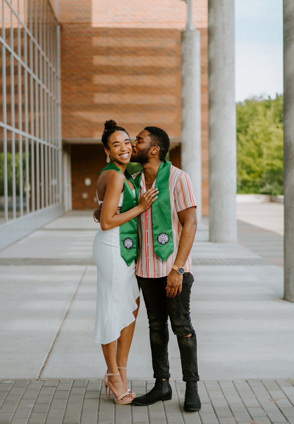 A couple wearing green graduation stoles embraces on a concrete walkway outside a modern building.