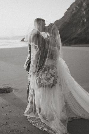 A romantic black and white beach wedding photo with a veiled couple standing on the sand near mountains.