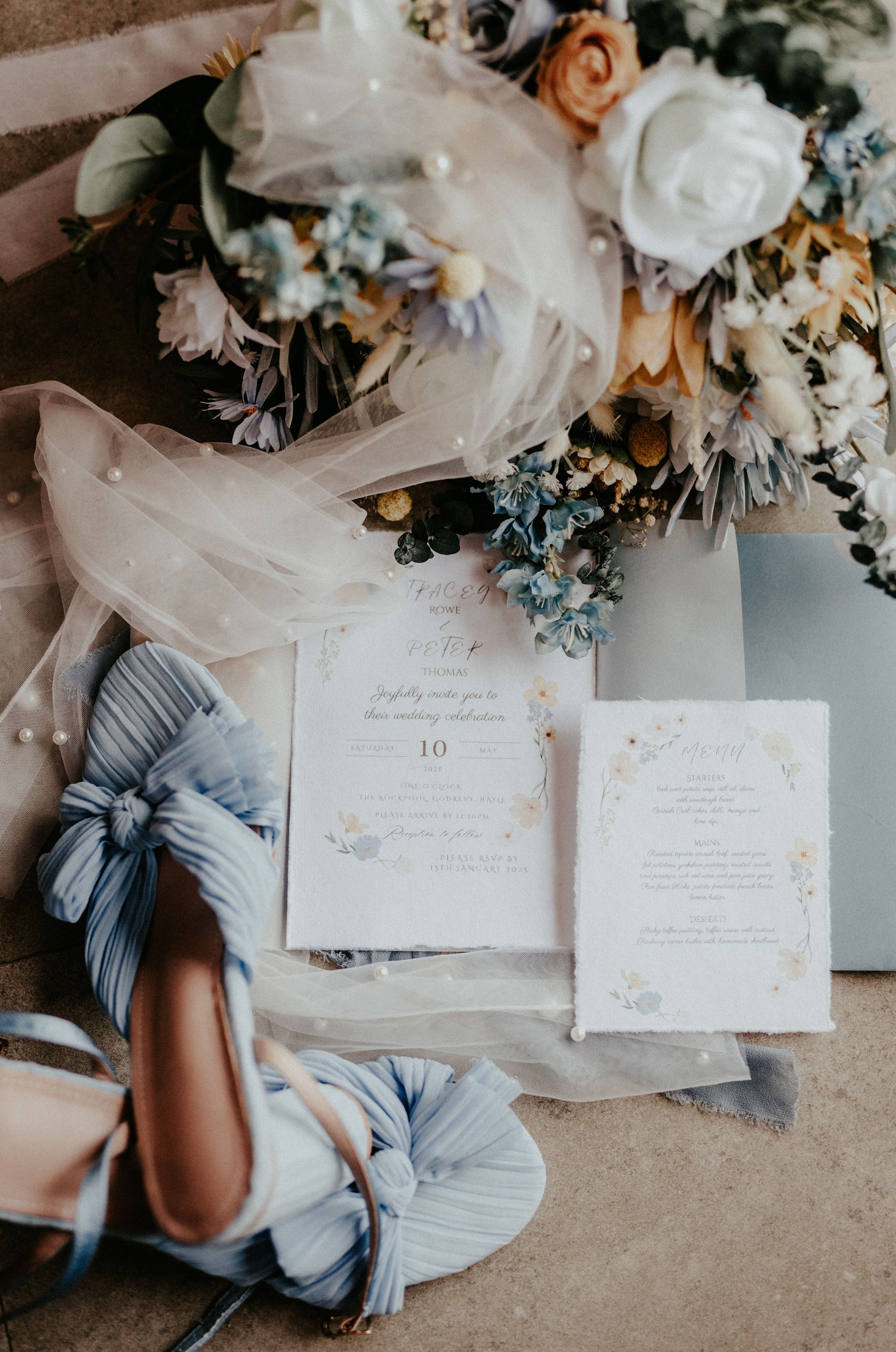 Elegant wedding invitation cards with light blue shoes and delicate floral arrangements on textured fabric background.