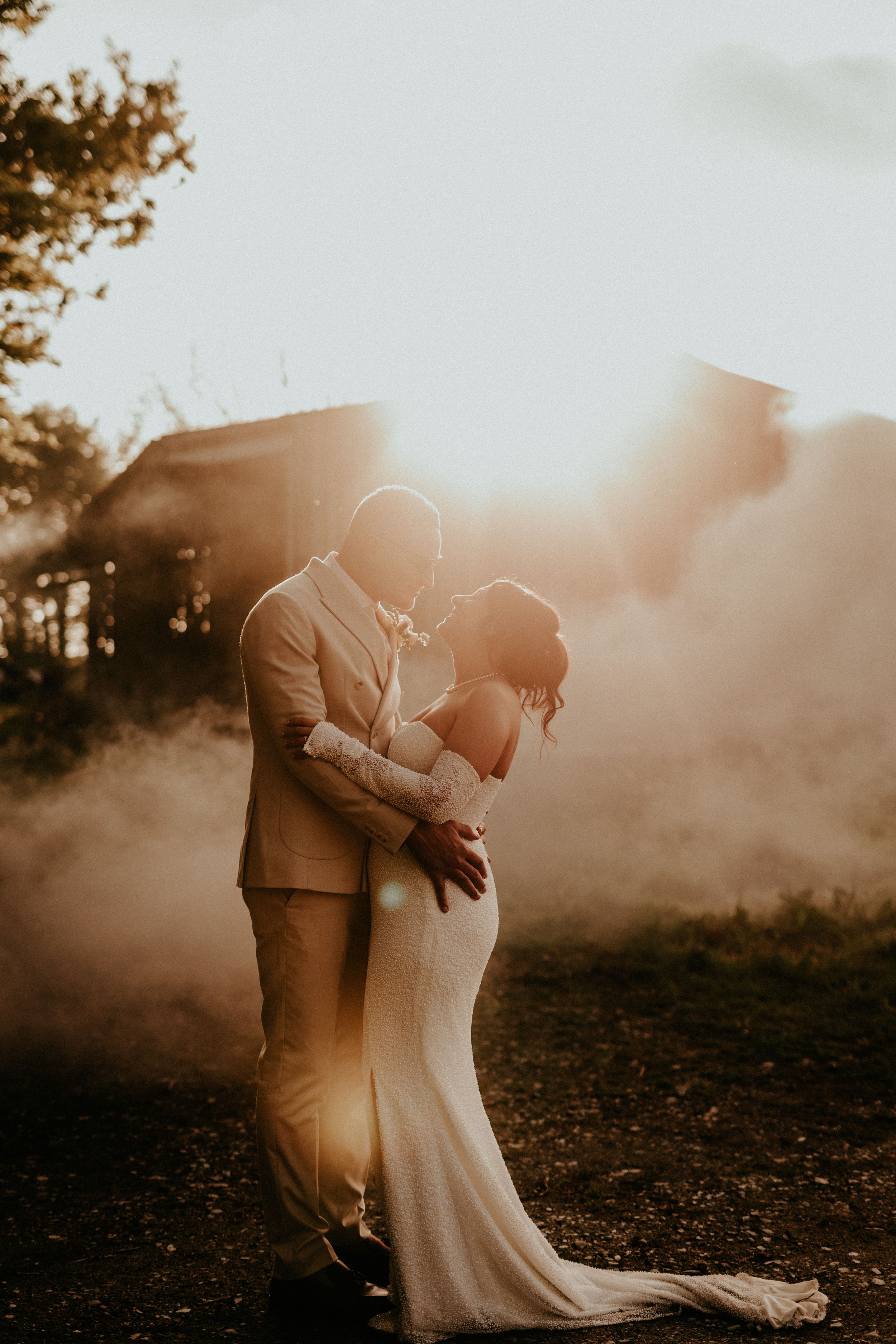 A romantic embrace between a couple on their wedding day is captured in ethereal golden sunlight and atmospheric mist.