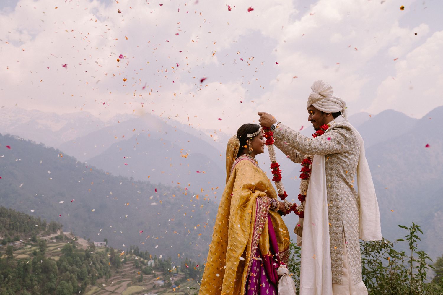 A traditional Indian wedding ceremony takes place in the mountains with flower petals falling through the air.