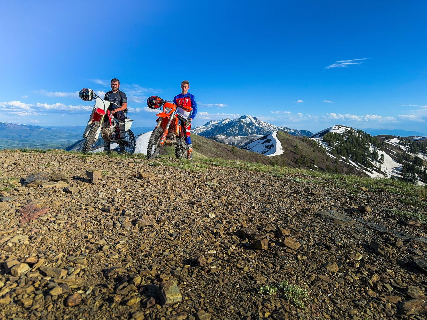 Two dirt bikers stand on a rocky mountain peak with snow-capped mountains and blue sky in the background.