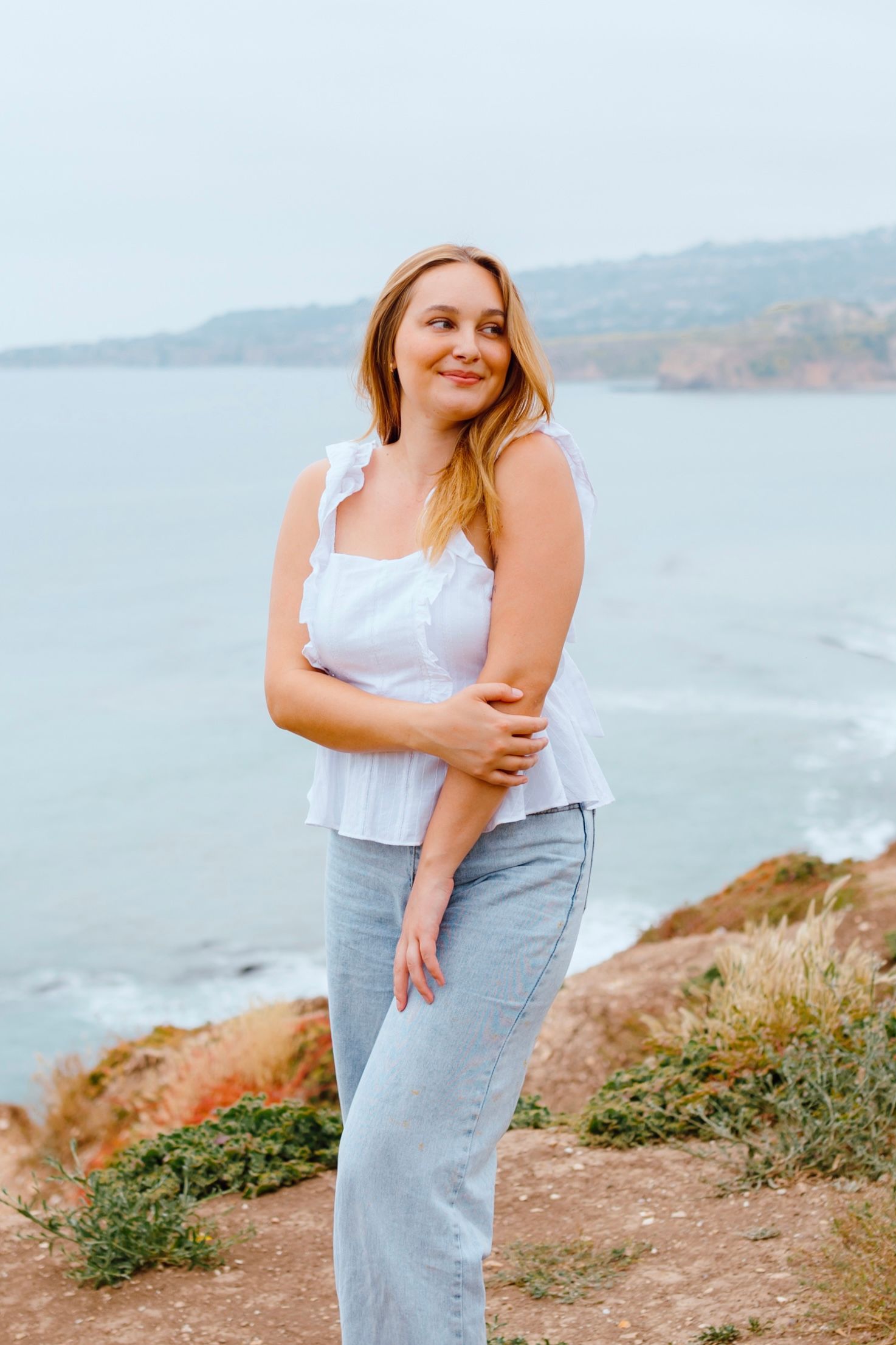 Photographer standing on coastal cliffs wearing light tank top and jeans with ocean view.
