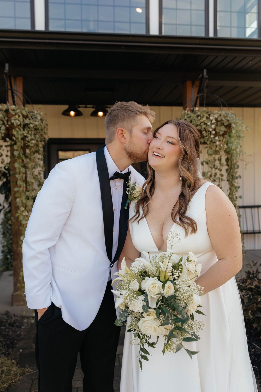 Romantic wedding couple shares intimate moments in front of modern venue with beautiful white floral bouquet.