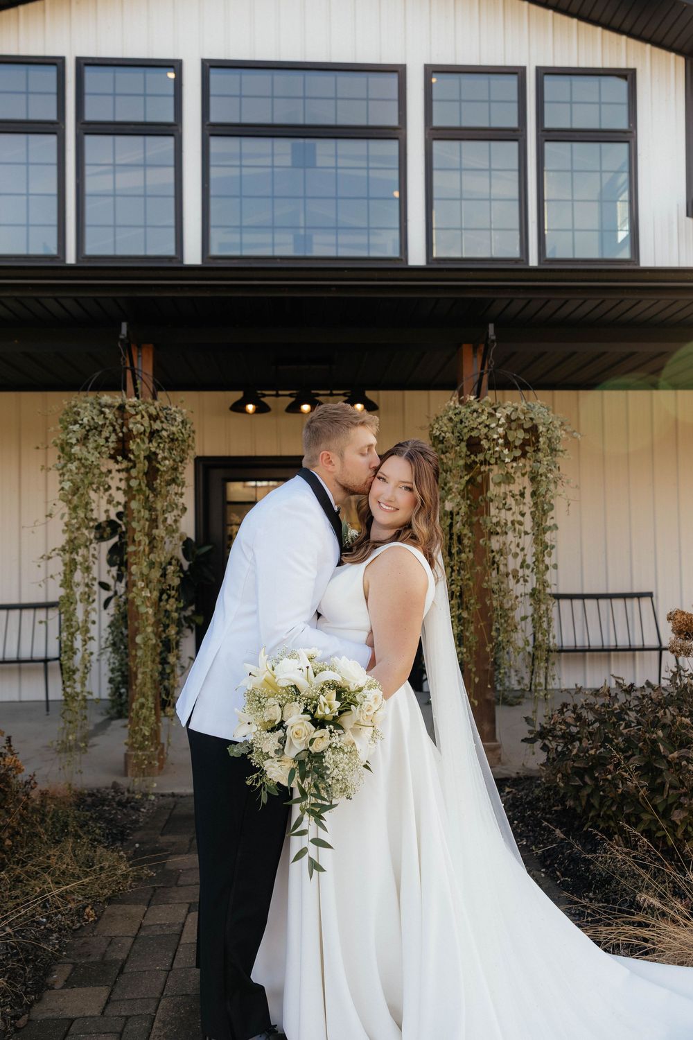 Modern white farmhouse wedding venue with large windows and climbing vines on the exterior columns.