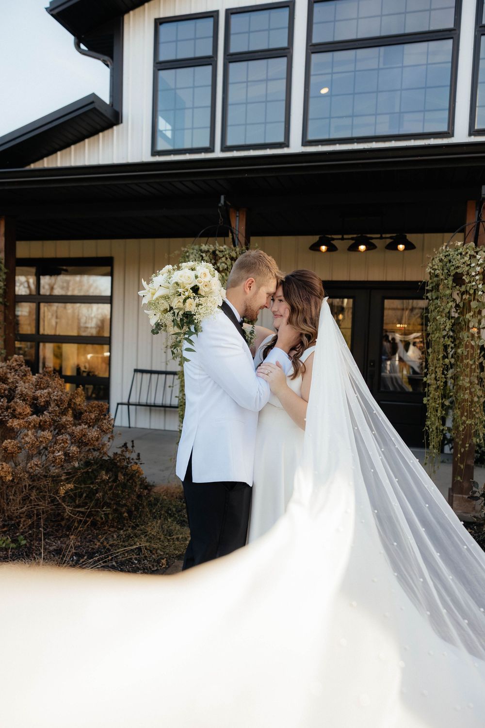A romantic wedding couple shares a tender moment outside a modern venue with a flowing white veil in the breeze.