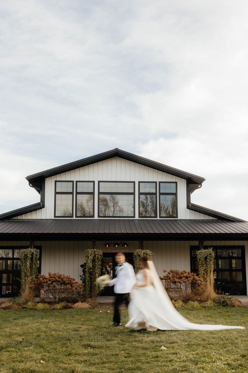 White farmhouse-style wedding venue with black trim and large windows under an overcast sky.