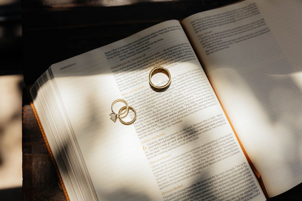 Two gold wedding rings resting on an open Bible, bathed in soft natural light and shadow.