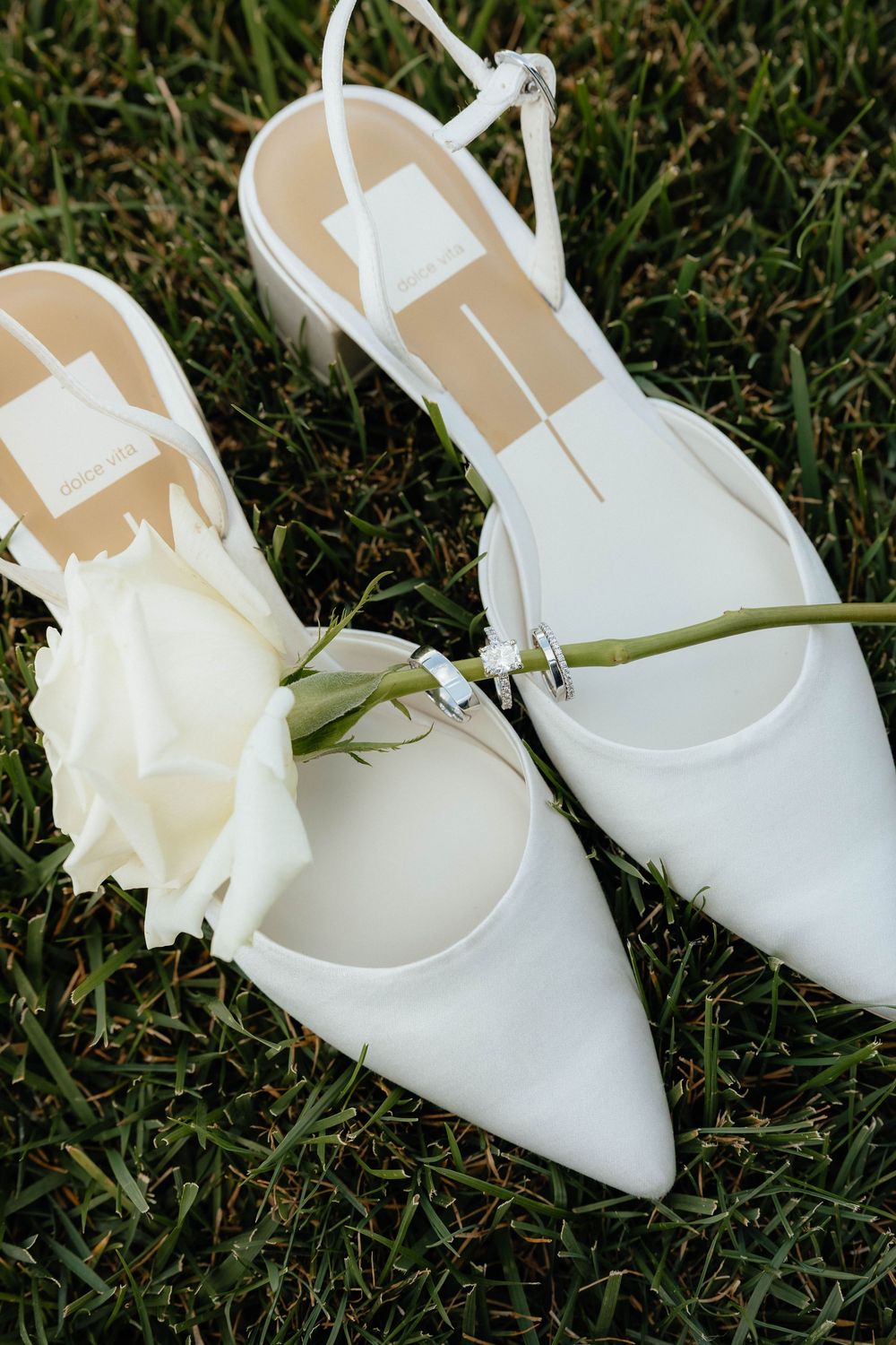 White slingback heels with pointed toes resting on green grass, adorned with a delicate white rose for a bridal aesthetic.