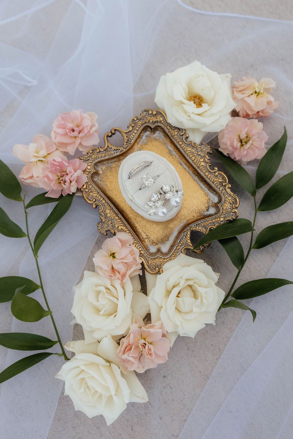 Ornate gold frame with wedding rings surrounded by cream and pink roses on white tulle.