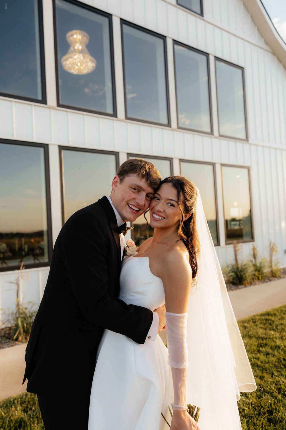 Smiling bride and groom embrace outside a white barn venue with a chandelier visible through windows.