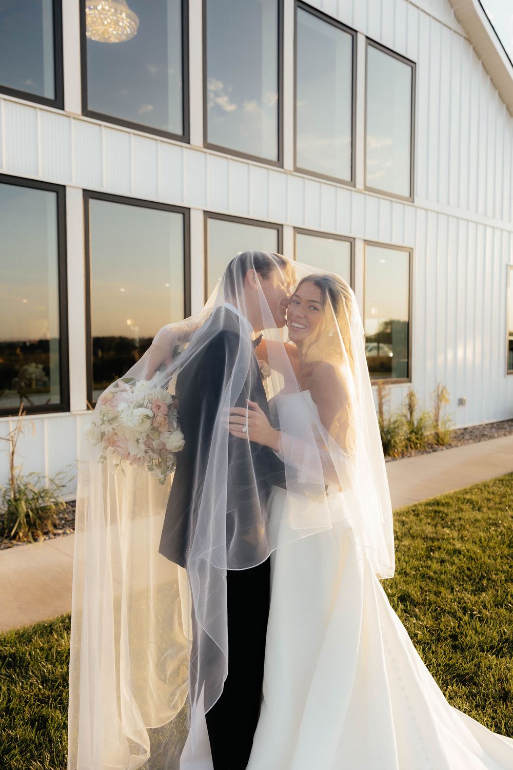 Bride and groom sharing a kiss under a flowing veil outside a modern white barn wedding venue at golden hour.