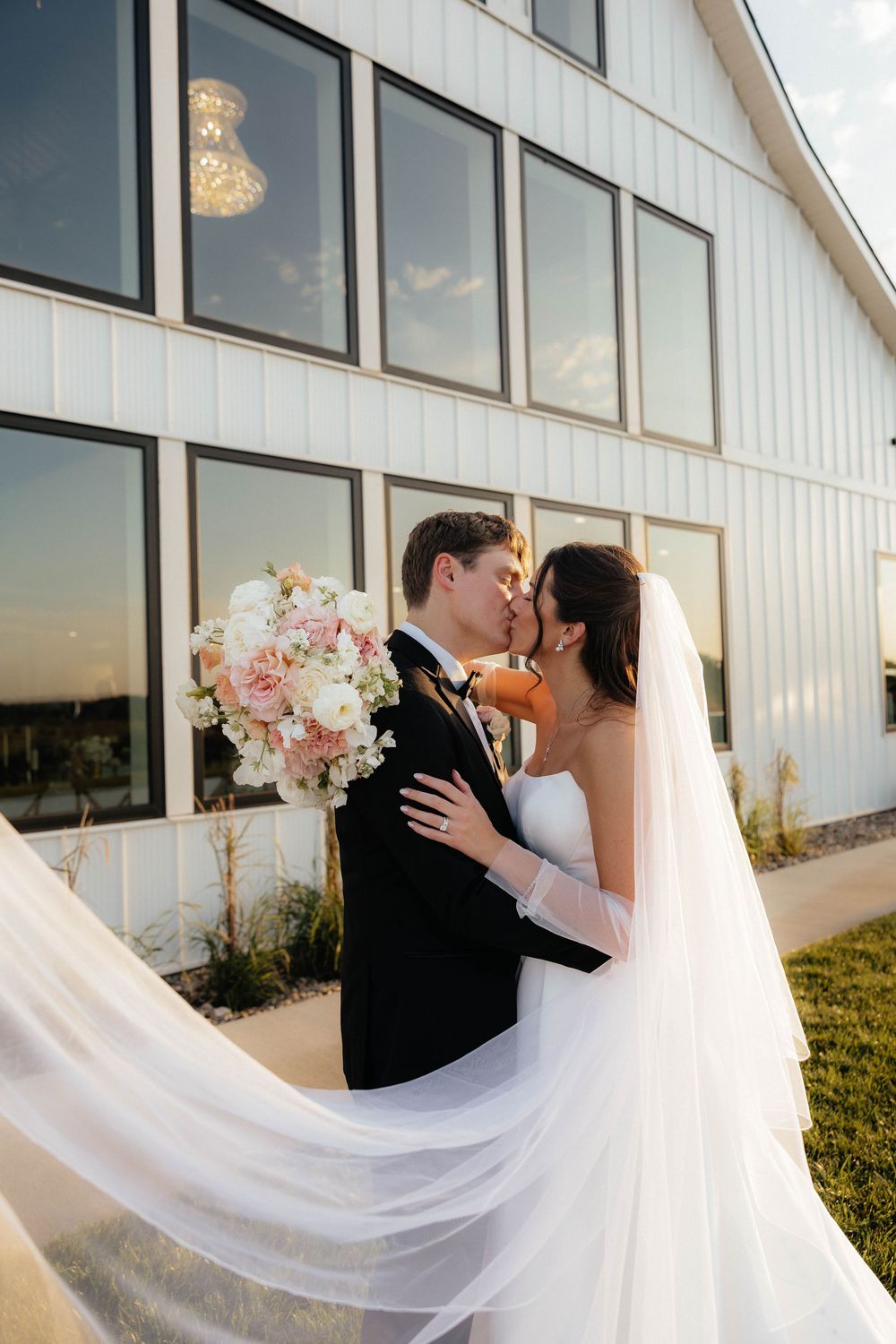 Bride and groom share a romantic kiss outdoors, with a flowing veil and blush bouquet in front of a white barn venue.