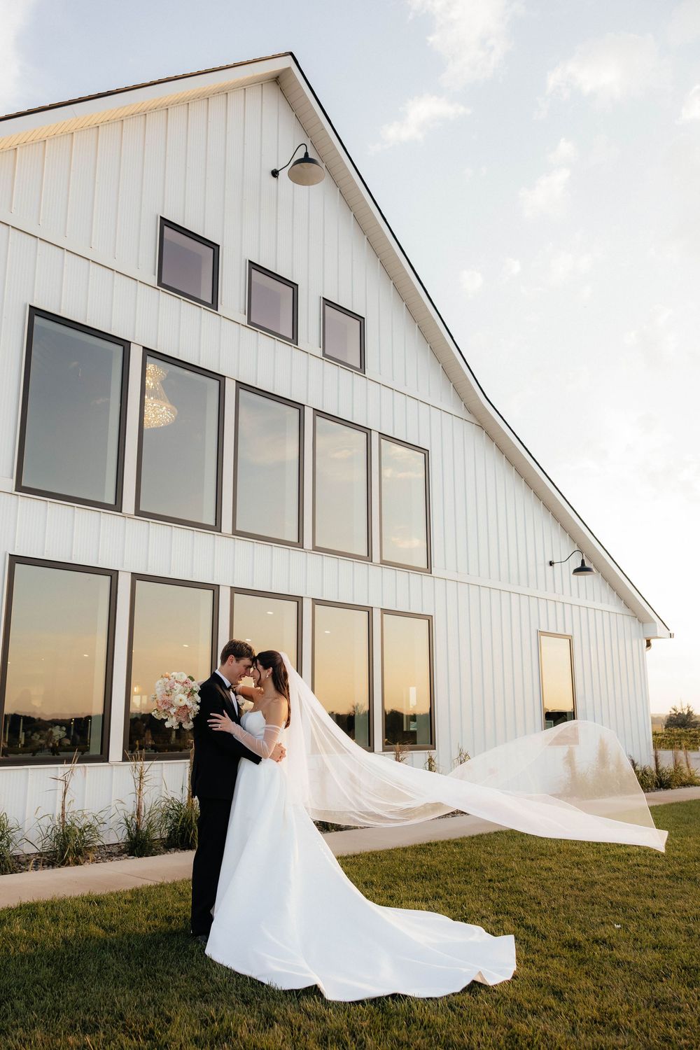Bride and groom embrace outdoors at golden hour before a modern white barn wedding venue, veil flowing in the wind.