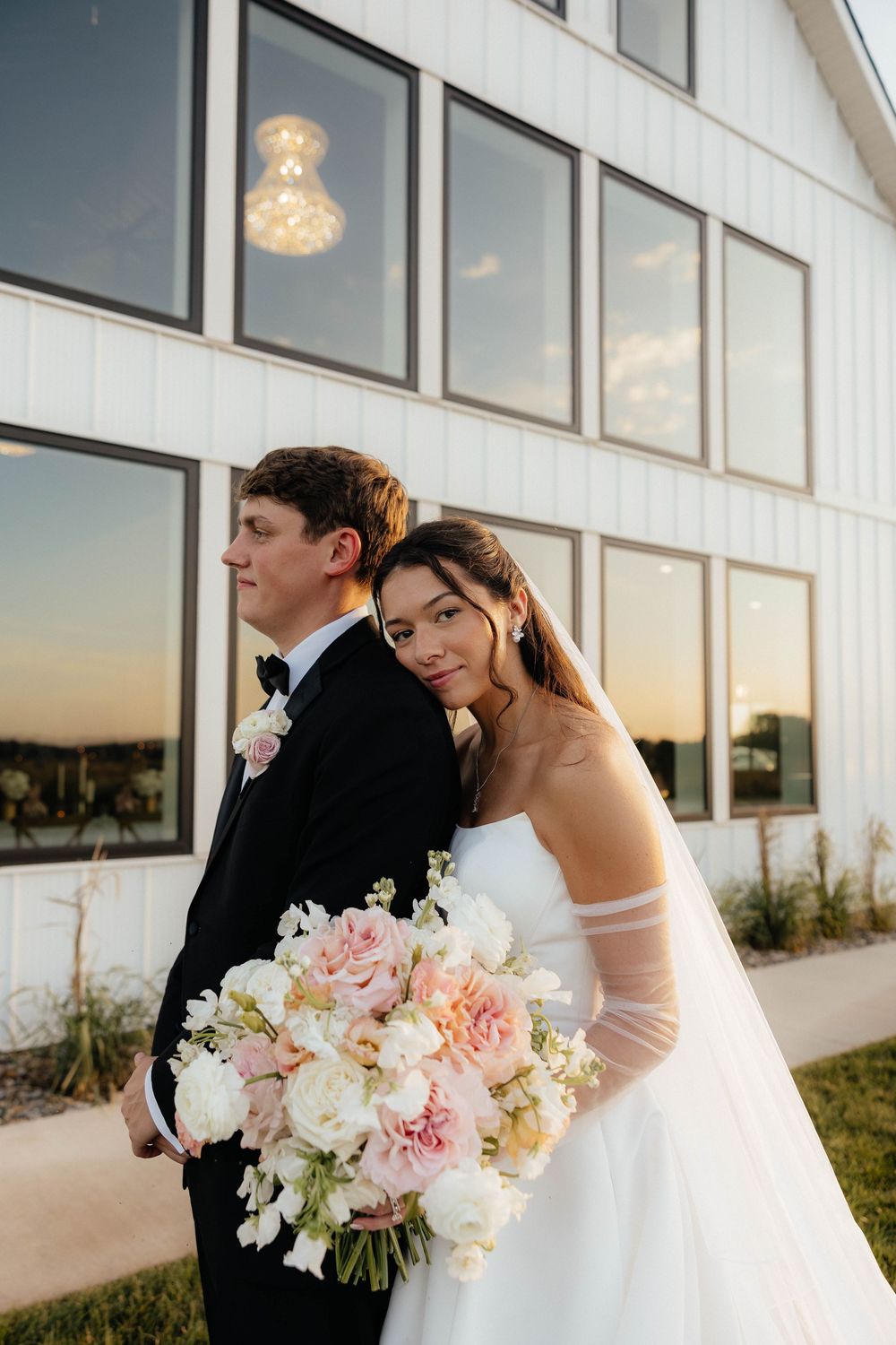 Bride leans against groom holding a blush and white peony bouquet outside a modern white barn venue at sunset.
