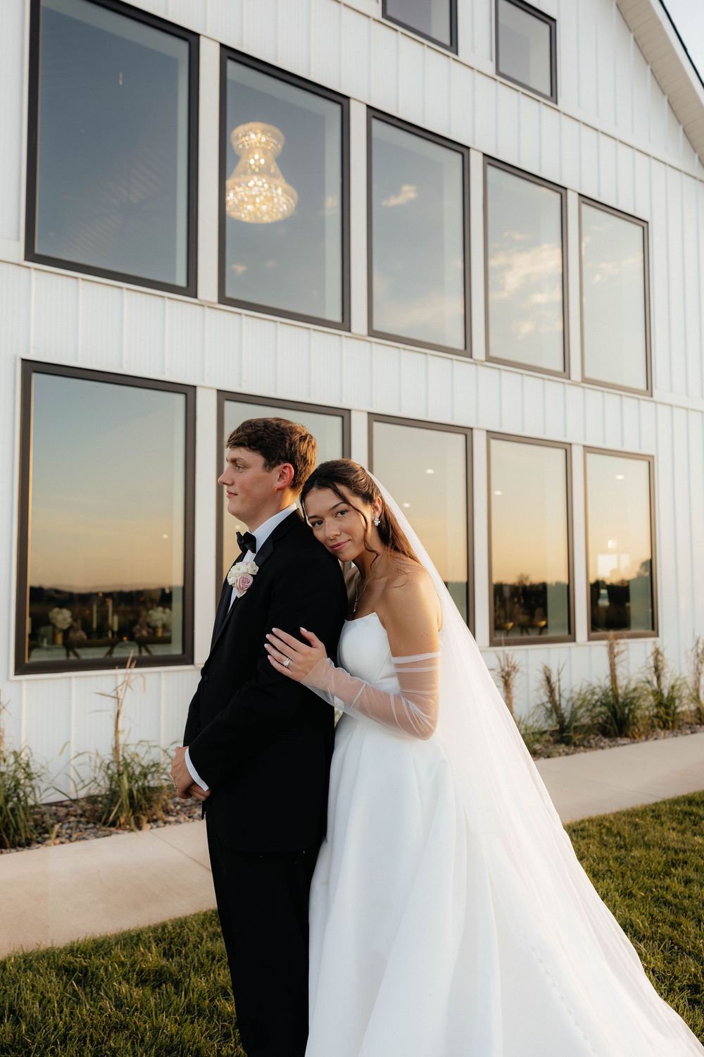 Bride in white gown leans on groom in black tux outside modern white venue at sunset.