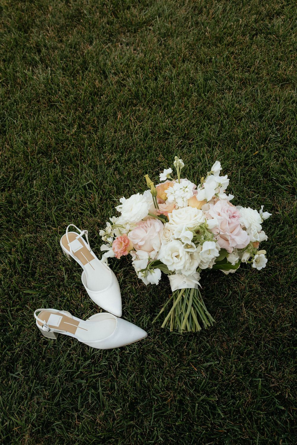 White bridal heels and a pastel pink and white wedding bouquet resting on lush green grass.