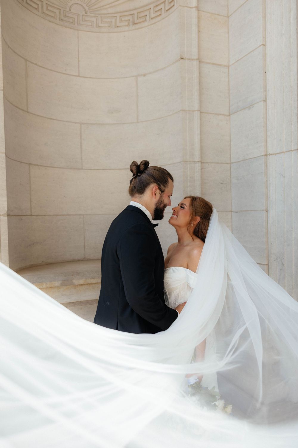 A bride and groom share a romantic moment, her flowing veil billowing, against an elegant stone column backdrop.