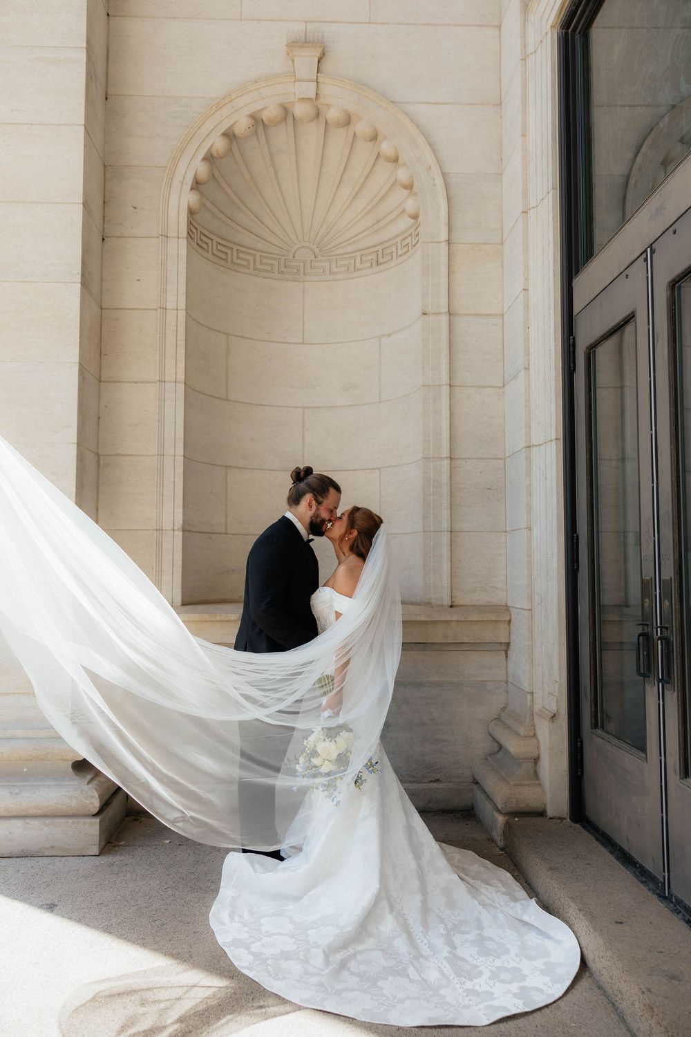 Bride and groom kissing beneath a flowing veil outside an elegant stone building on their wedding day.