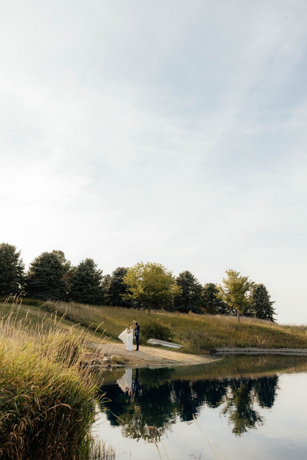 A scenic lakeside wedding photo with tall grass and trees reflecting in calm waters at sunset.