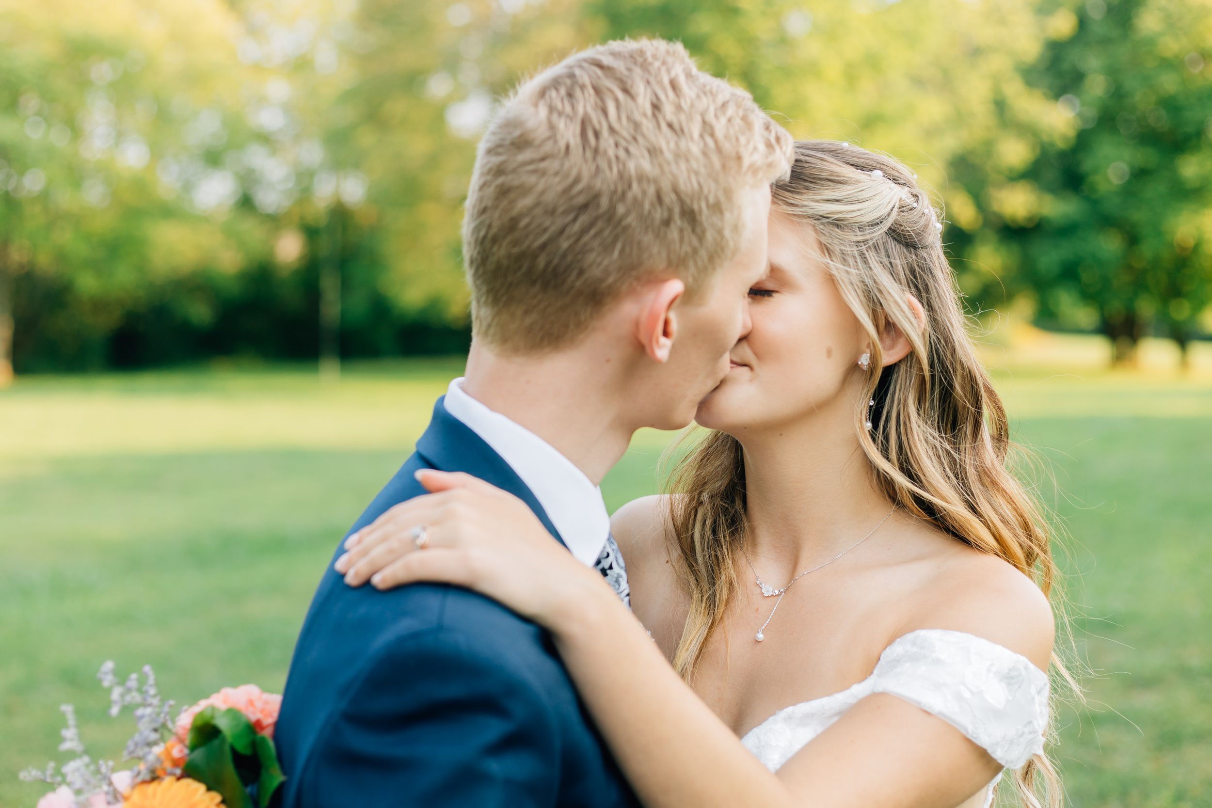 A couple shares a romantic kiss in formal wedding attire on a sunny grass field.