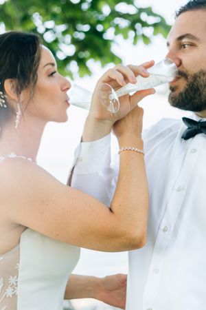 Wedding couple sharing a celebratory toast outdoors in bright natural lighting.