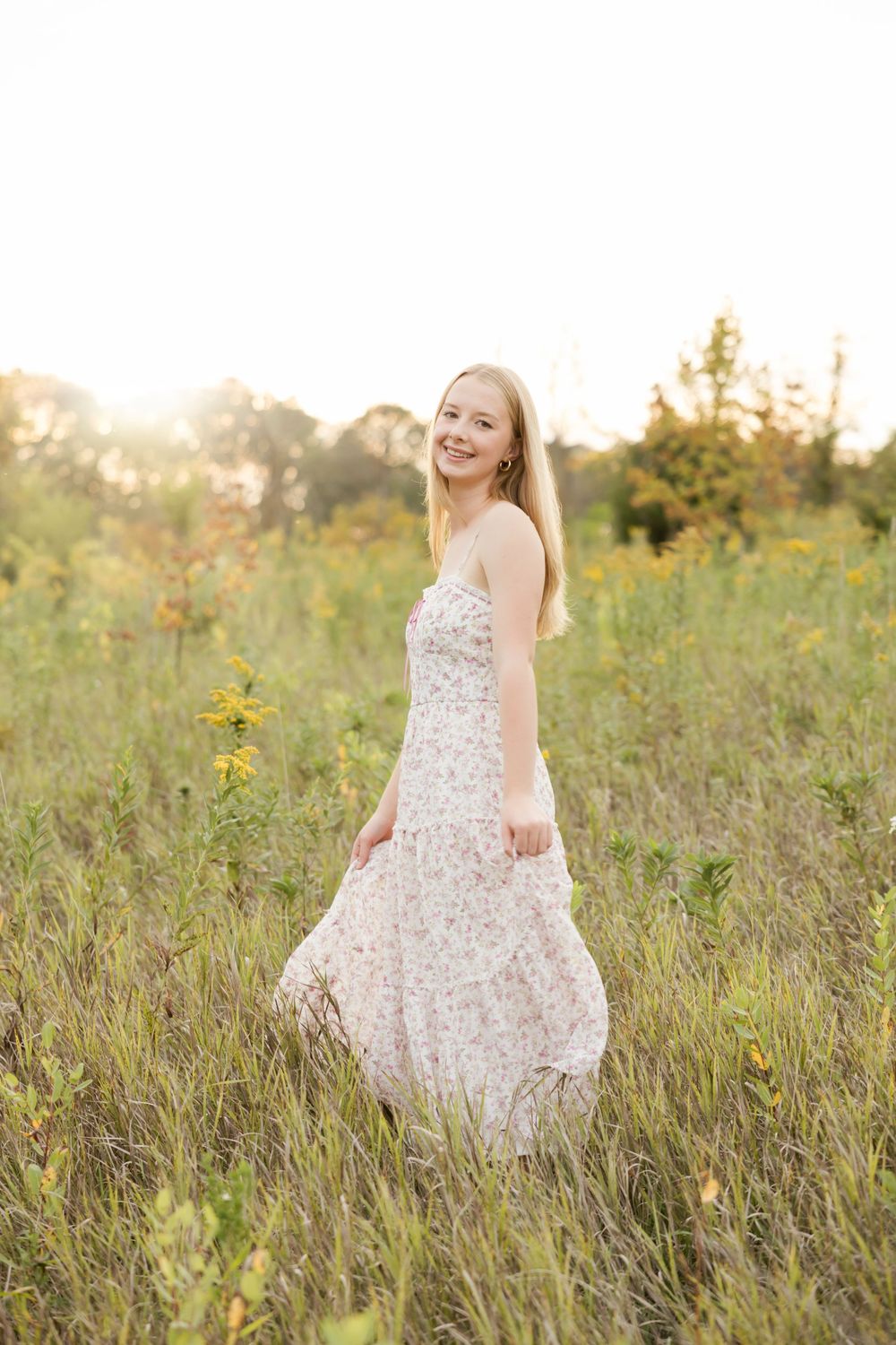 A person in a floral summer dress stands in a sunlit meadow with golden wildflowers during sunset.