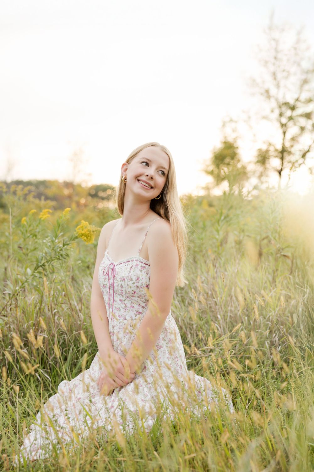 A person in a floral sundress sits in a sunlit meadow with wildflowers during golden hour.
