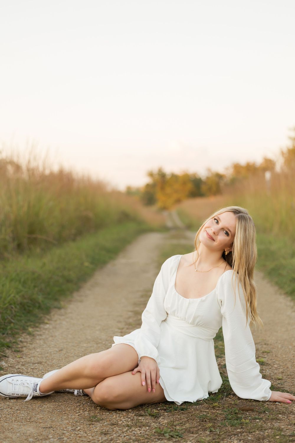 A person in a flowing white dress poses gracefully on a dirt path during golden hour.