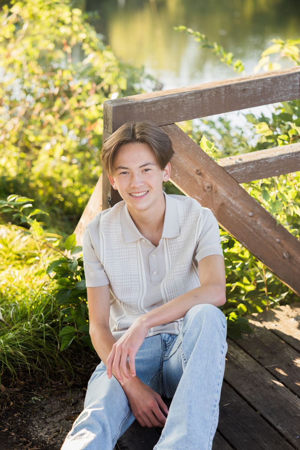 Person in white button-down shirt and jeans sits casually on wooden deck surrounded by lush greenery in natural light.