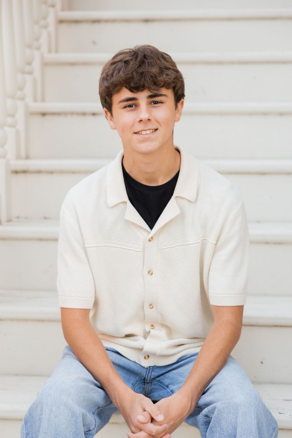 A person in a white polo shirt and blue jeans sits casually on white steps in a bright outdoor setting.