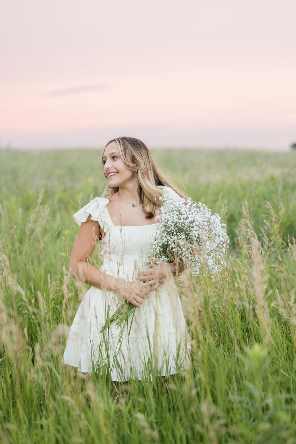 Person in white dress standing among tall grass with baby's breath bouquet at sunset.