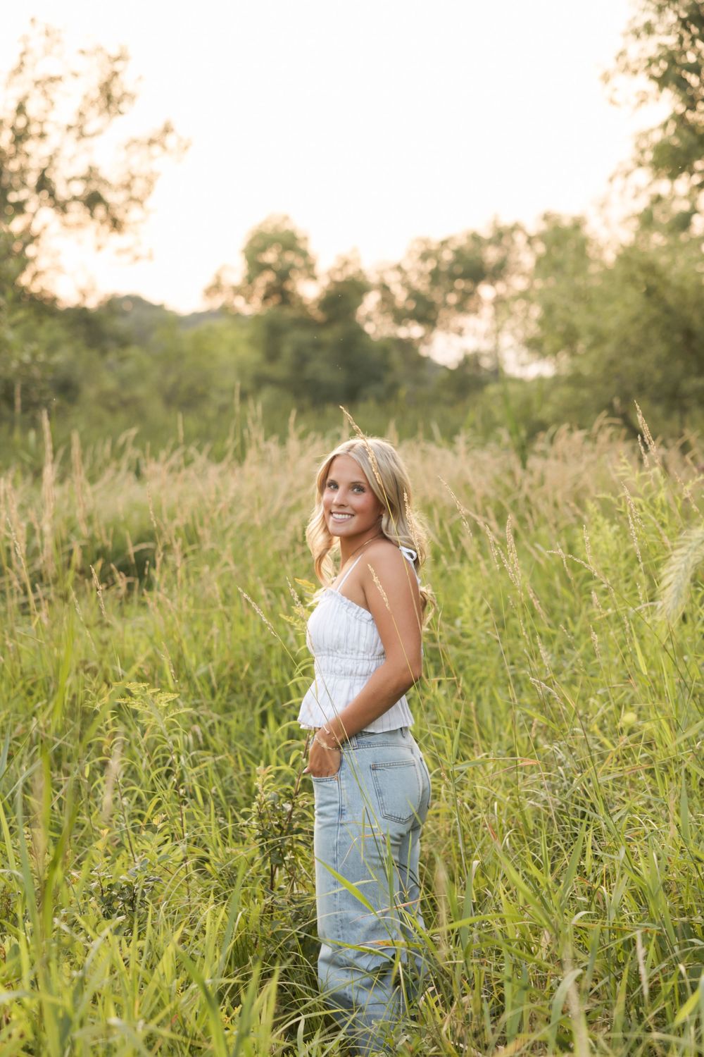 Smiling blonde woman in white top and jeans standing in tall green grass field during golden hour sunset.