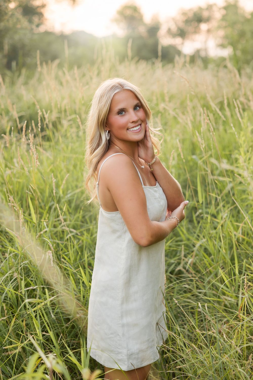 Smiling blonde woman in white sundress standing in tall green grass field during golden hour sunset.