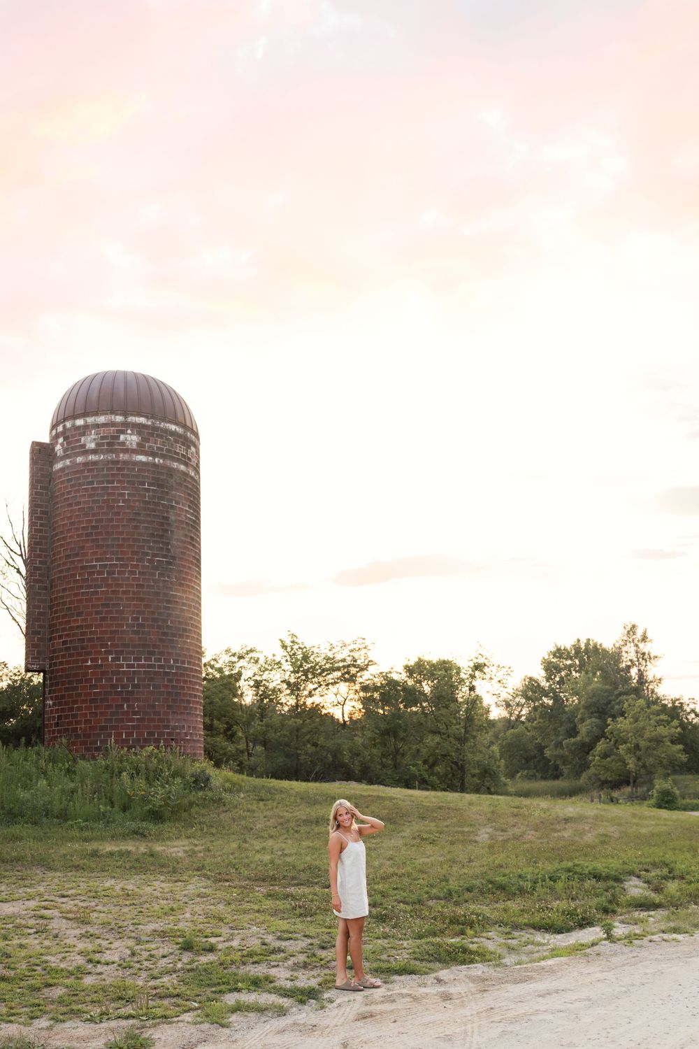 Young girl in white dress stands beside a tall brick silo at sunset on a rural farm.