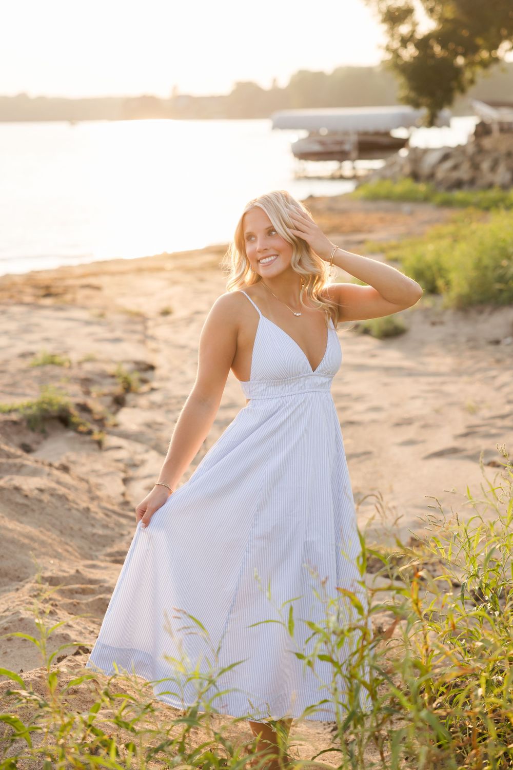Smiling blonde woman in white sundress posing on sandy beach at golden hour sunset near waterfront.