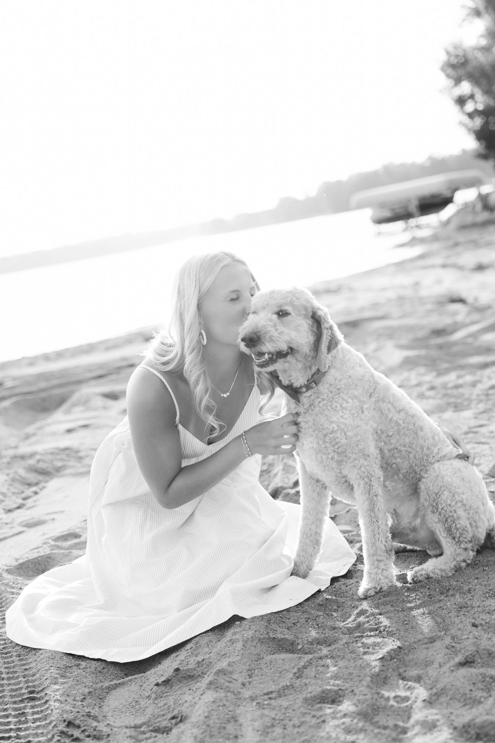 Black and white photo of a blonde woman in a white dress kissing a golden doodle dog on a sandy beach.