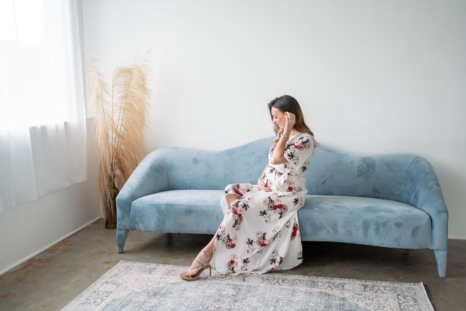 Woman in floral maxi dress sits elegantly on light blue sofa with pampas grass decor in minimalist room.