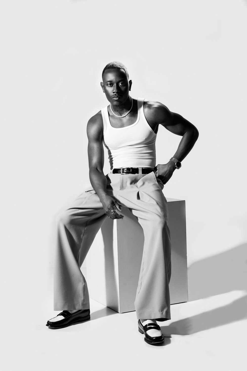 Black and white photo of a muscular man in a white tank top and wide-leg trousers sitting on a white cube.