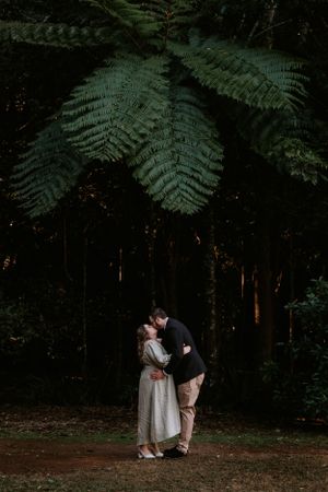 Couple embracing under large fern leaves in moody forest setting.
