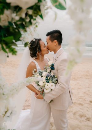 Romantic wedding couple embraces on a sandy beach with white floral arrangements and soft natural lighting.