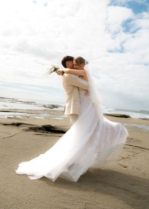 A couple in wedding attire dance and twirl together on a scenic beach with cloudy skies and rocky shoreline.