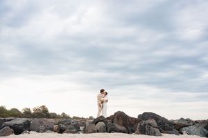 Wedding couple standing together on rocky coastal shoreline against cloudy sky during sunset photography session.