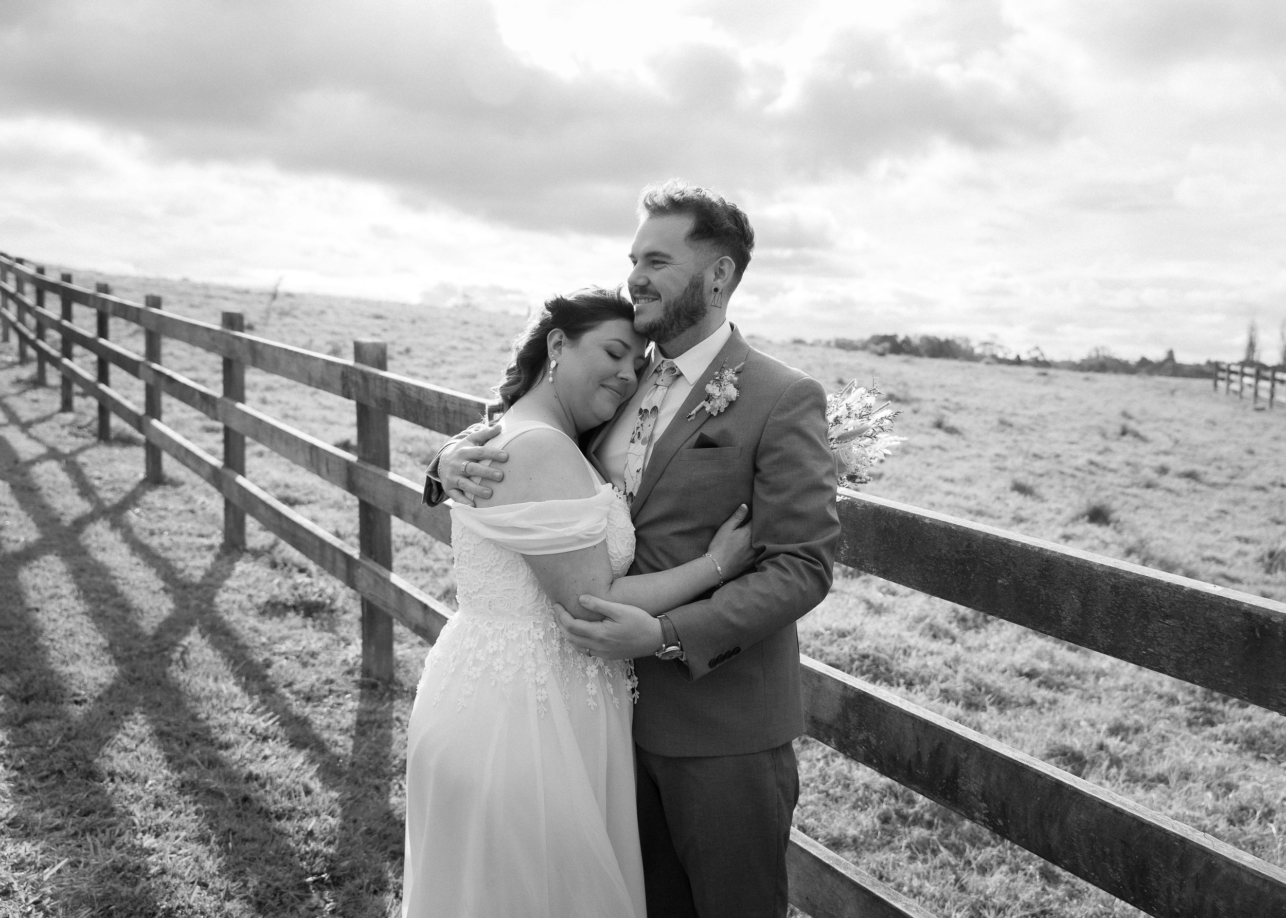 A couple in wedding attire embraces while dancing outdoors near a rustic wooden fence at sunset.