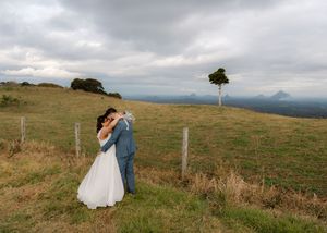 A couple in wedding attire embraces on a scenic hilltop meadow with mountains in the background.