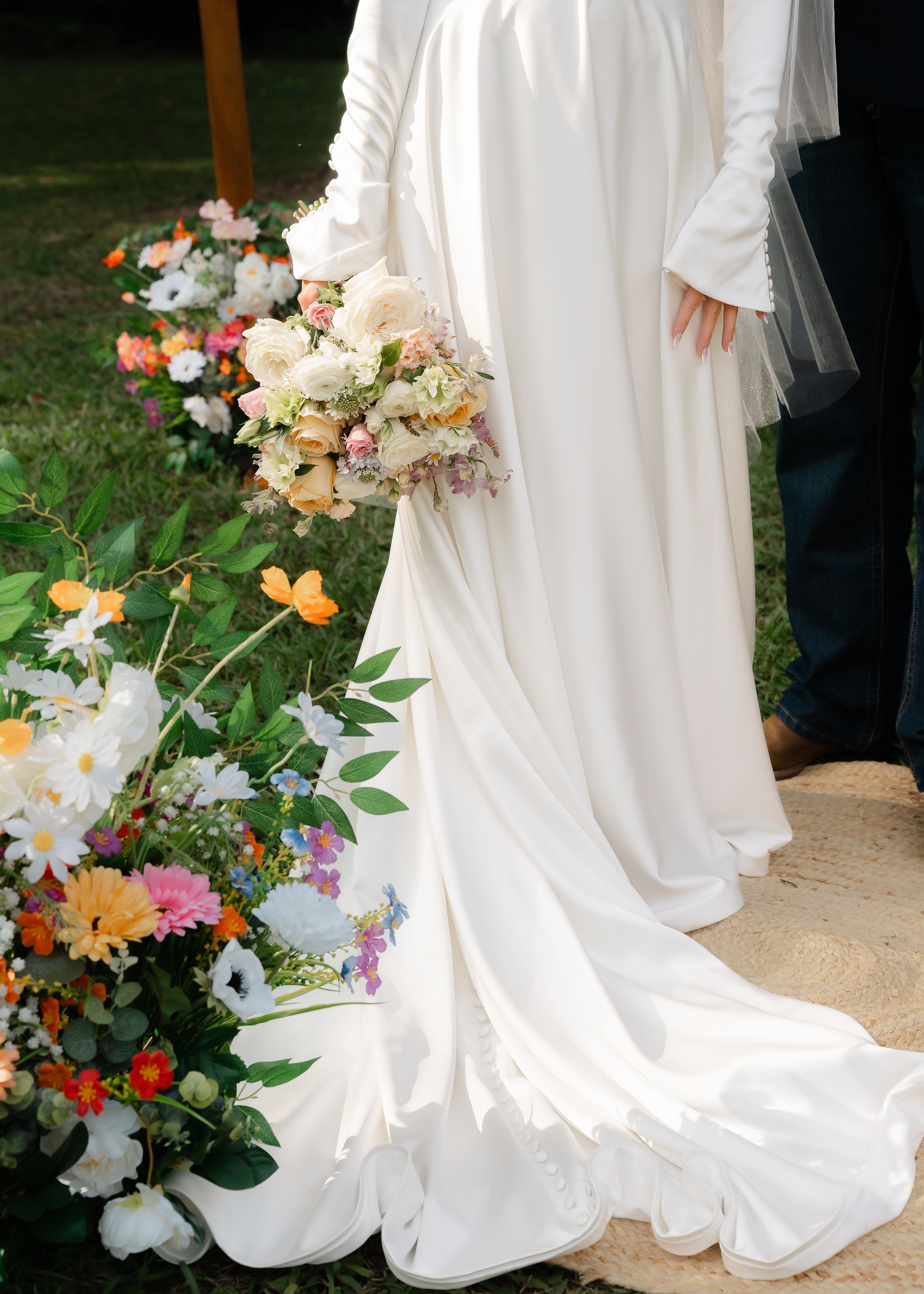 Elegant white wedding dress train flowing across stone pathway with colorful floral arrangements in garden setting.