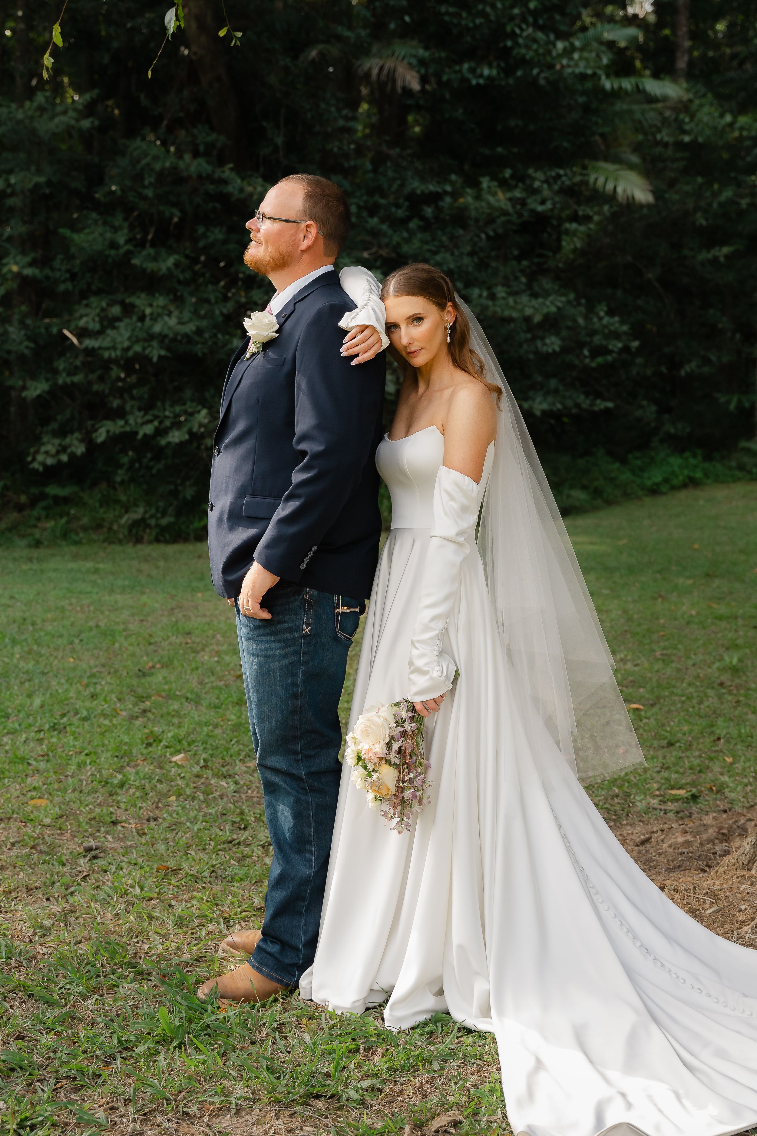 A bride in a flowing white gown with long train and veil stands with a companion in jeans and dress shirt outdoors.