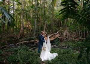 A couple in wedding attire shares an intimate moment surrounded by lush forest greenery and tall trees.