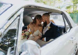 A couple shares an intimate moment in the backseat of a classic car while holding a wedding bouquet.